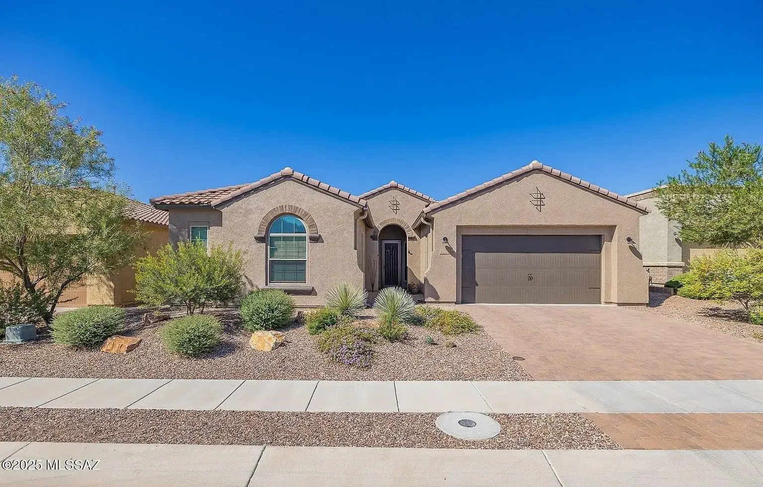 Single-story house with arched windows, desert landscaping, and a two-car garage under a clear blue sky.