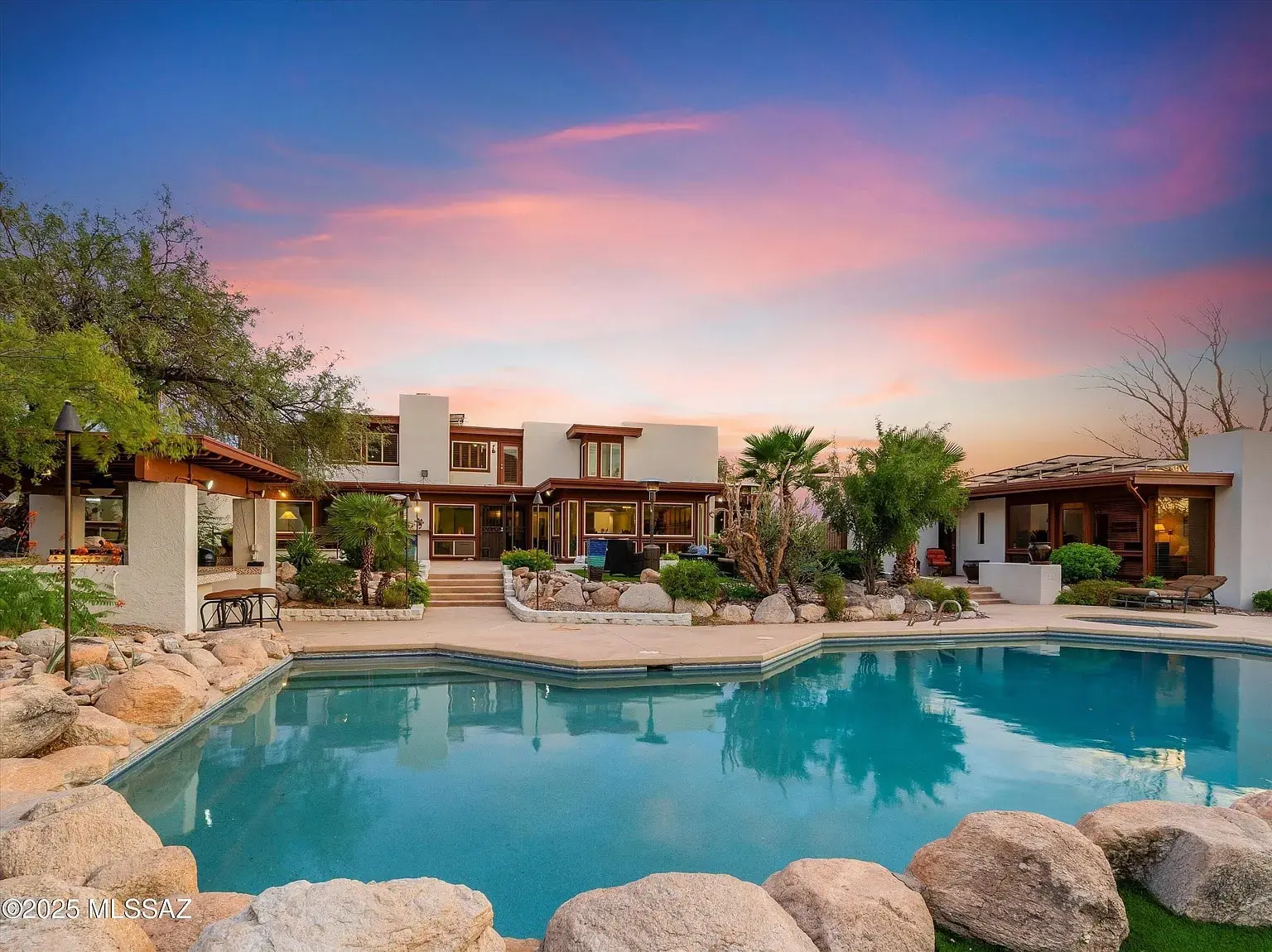 Luxury home with large pool, surrounded by rocks and greenery, under a vibrant sunset sky.