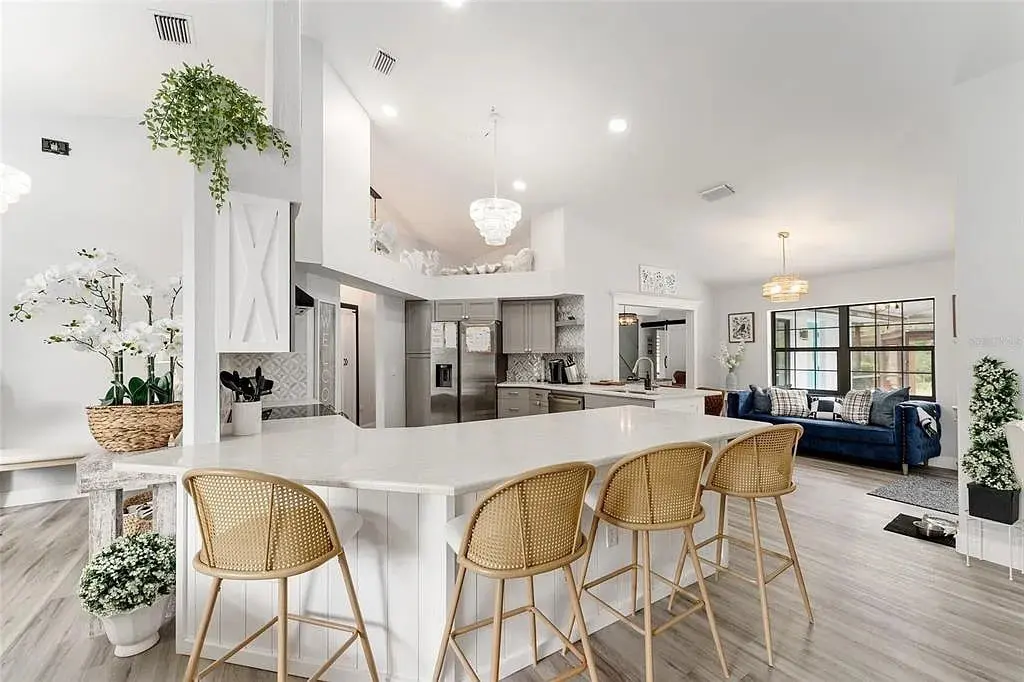 Modern kitchen and living area with white decor, barstools, plants, and a blue sofa.