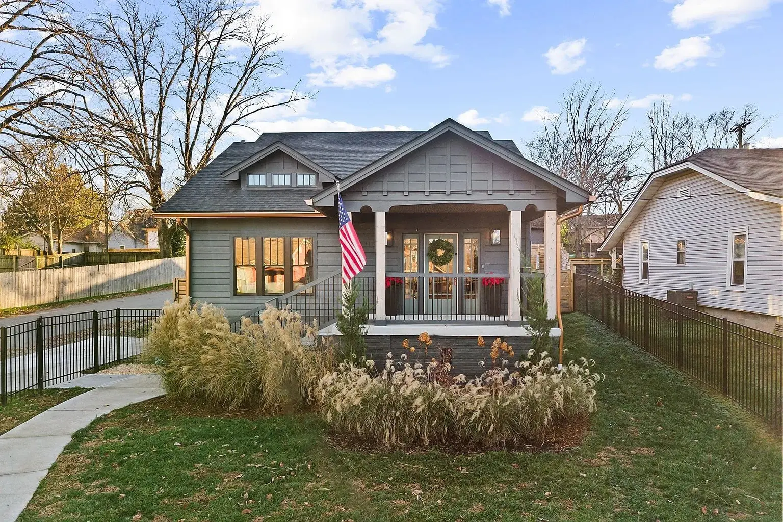 A cozy gray house with a front porch, American flag, and surrounding garden.