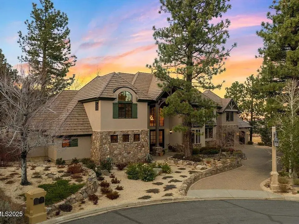 Large stone and stucco house surrounded by trees, with a sunset sky in the background.