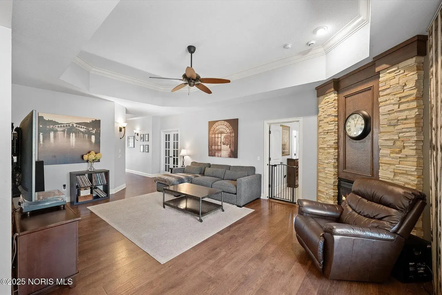 Spacious living room with gray sectional sofa, armchair, ceiling fan, and wall clock. Wood flooring.