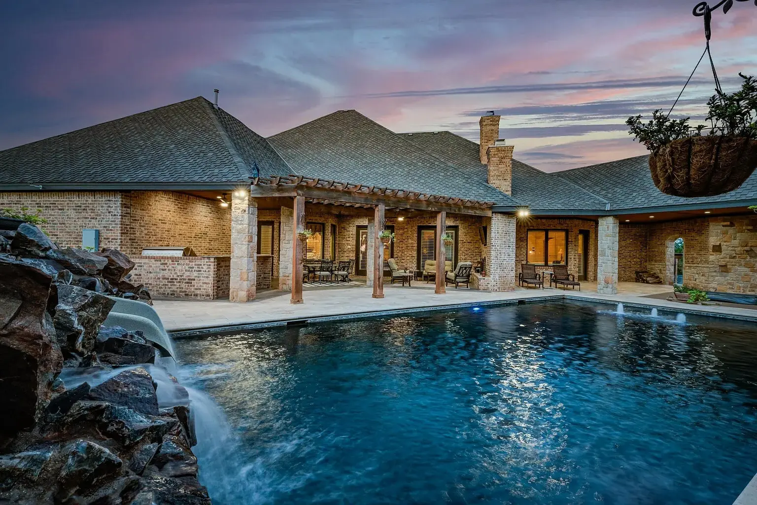 Large backyard pool with waterfall feature, surrounded by a brick house and patio at sunset.