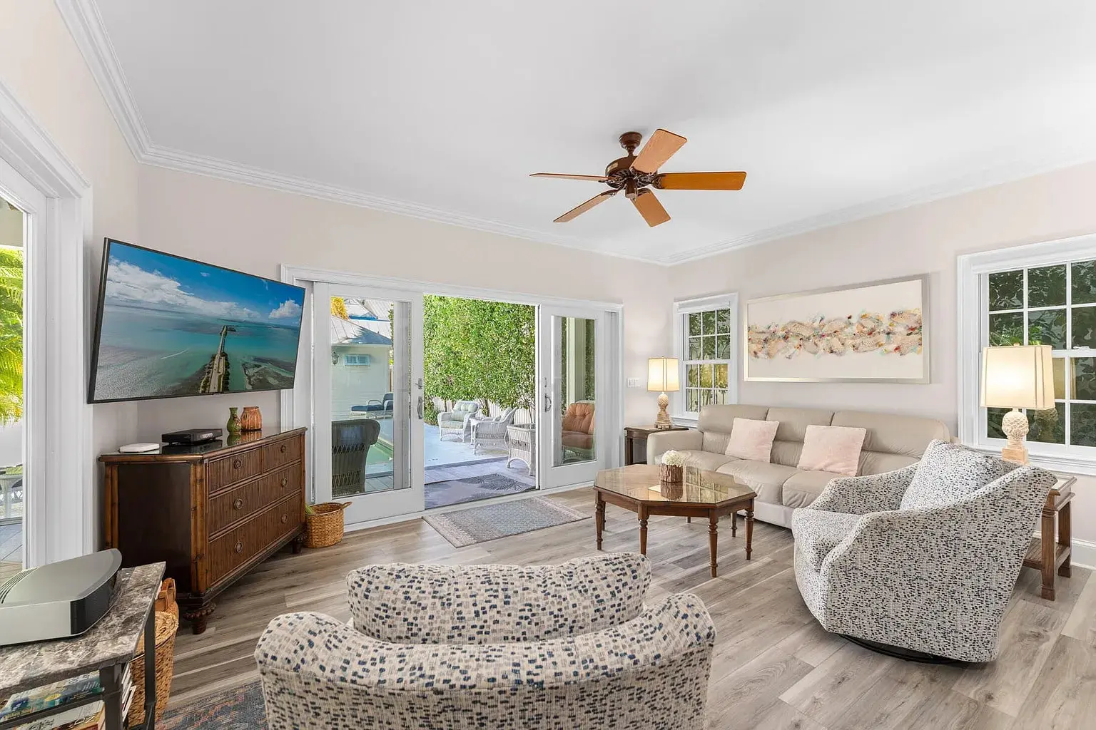 Bright living room with a ceiling fan, TV, sofa, armchairs, and glass doors leading outside.