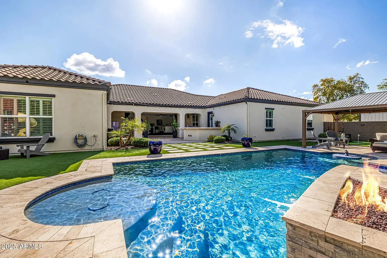 Backyard with a swimming pool, fire pit, and covered patio area under a sunny sky.