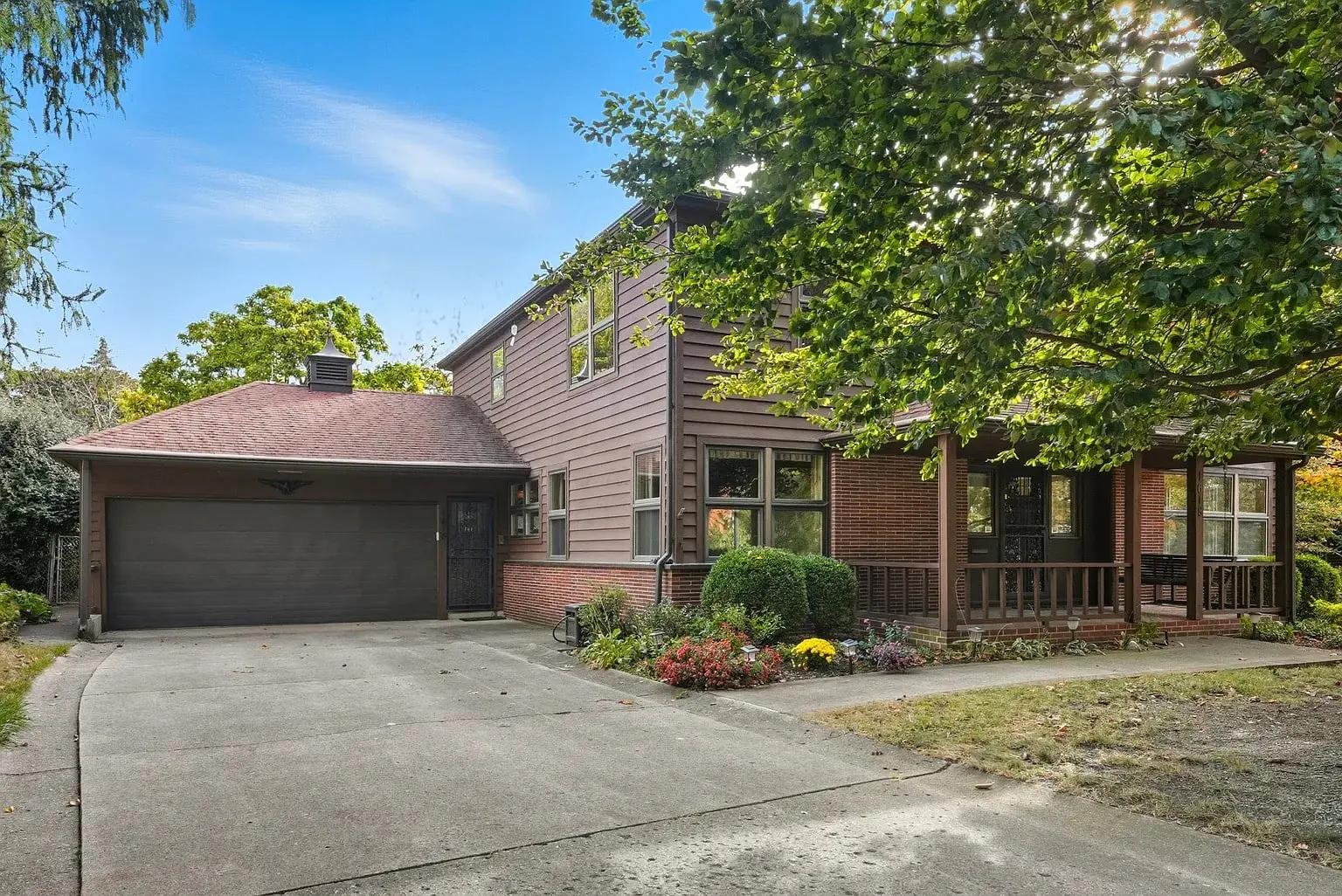 Two-story house with a garage, surrounded by trees and a garden, under a clear blue sky.