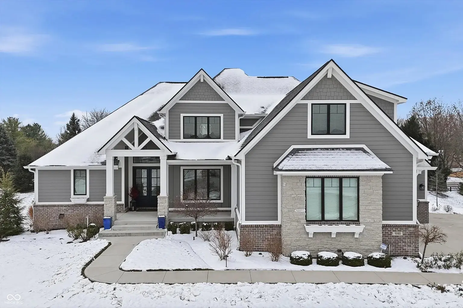 Gray two-story house with snow-covered roof and yard, featuring a front porch and driveway.
