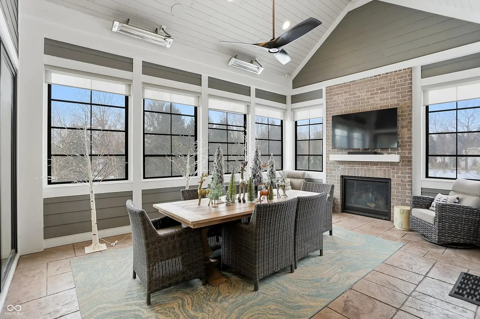 Sunroom with dining table, wicker chairs, fireplace, TV, and large windows overlooking snowy landscape.