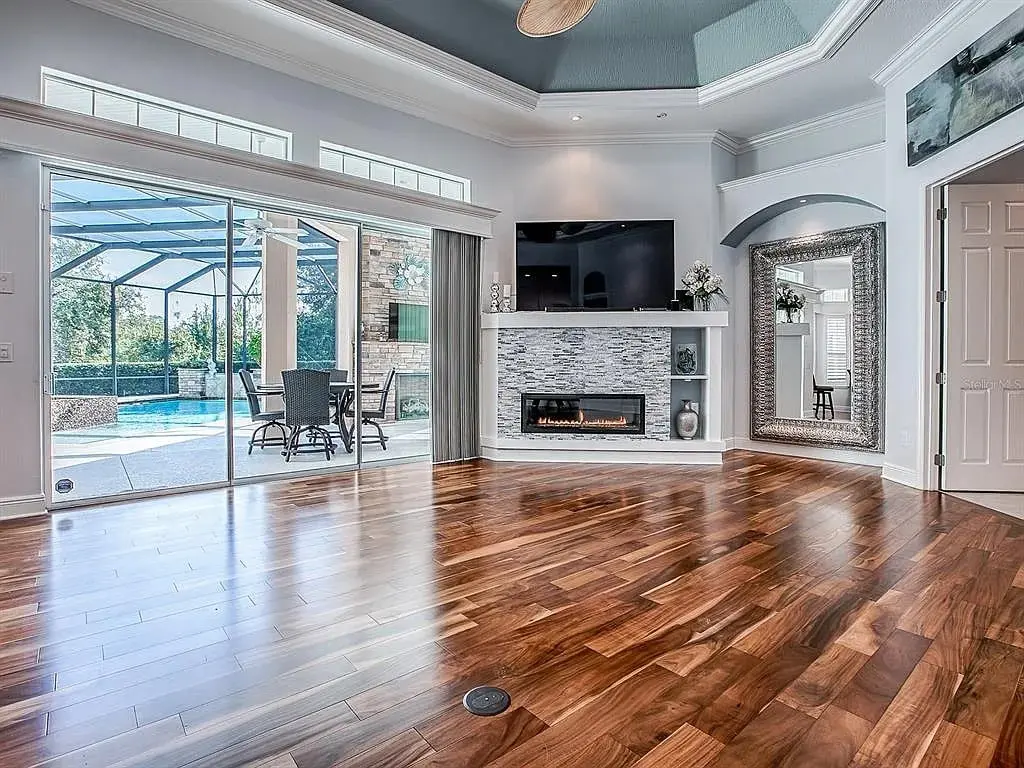 Spacious living room with wood flooring, fireplace, large TV, and glass doors leading to a pool.