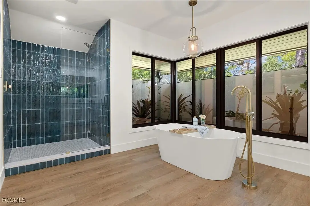Modern bathroom with freestanding bathtub, glass-enclosed shower, large windows, and wood flooring.