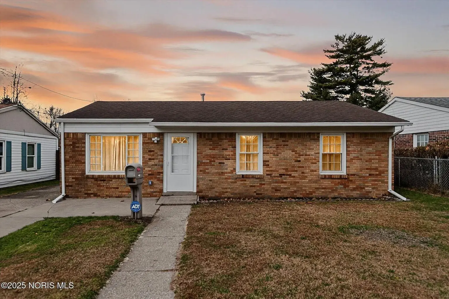 Single-story brick house with lit windows, a front lawn, and a sunset sky backdrop.