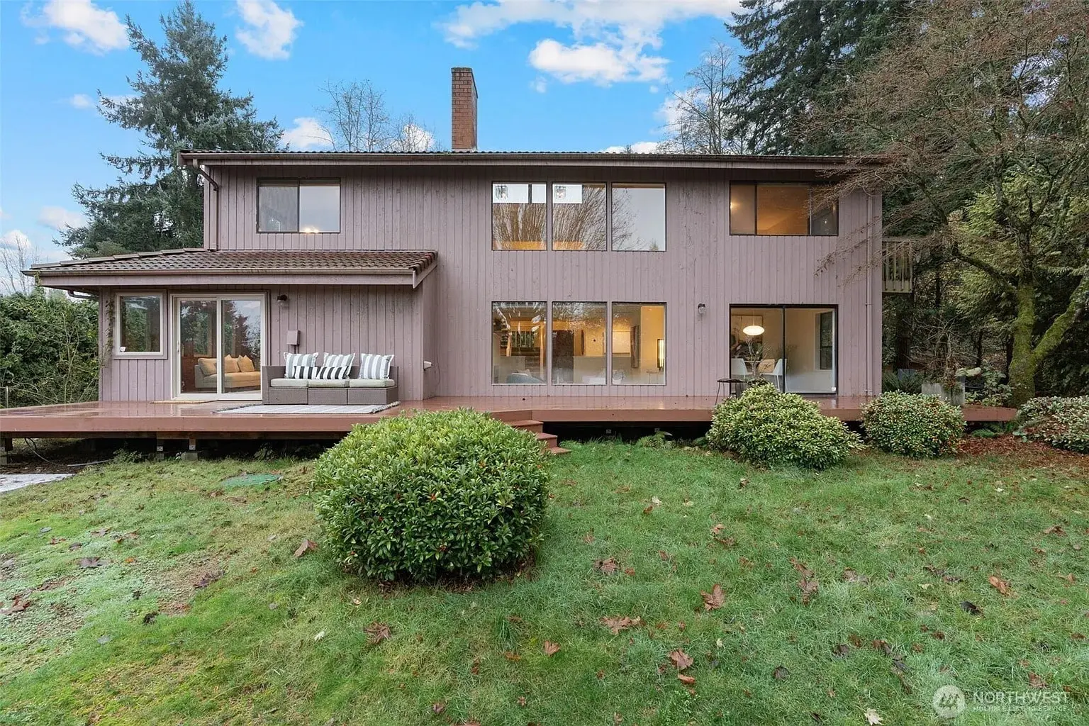 Two-story house with large windows, wooden deck, and surrounding greenery.