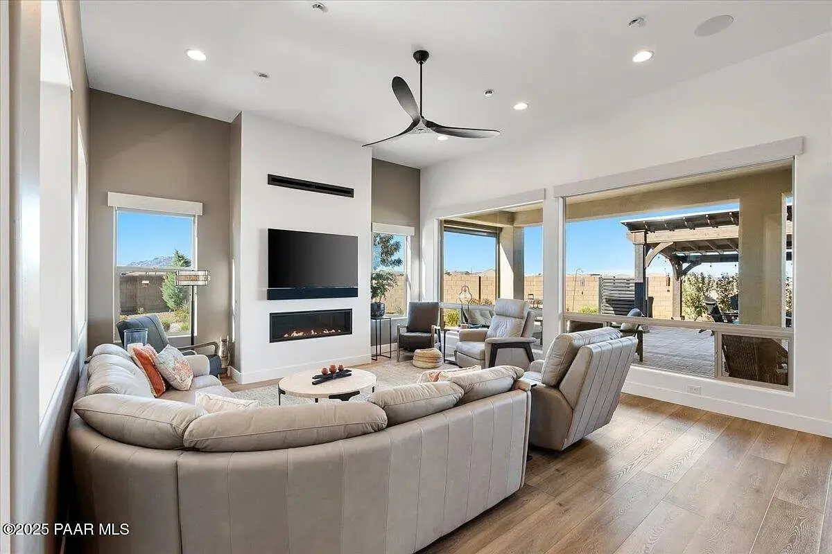 Modern living room with beige sectional sofa, recliners, wall-mounted TV, and large windows overlooking a patio.