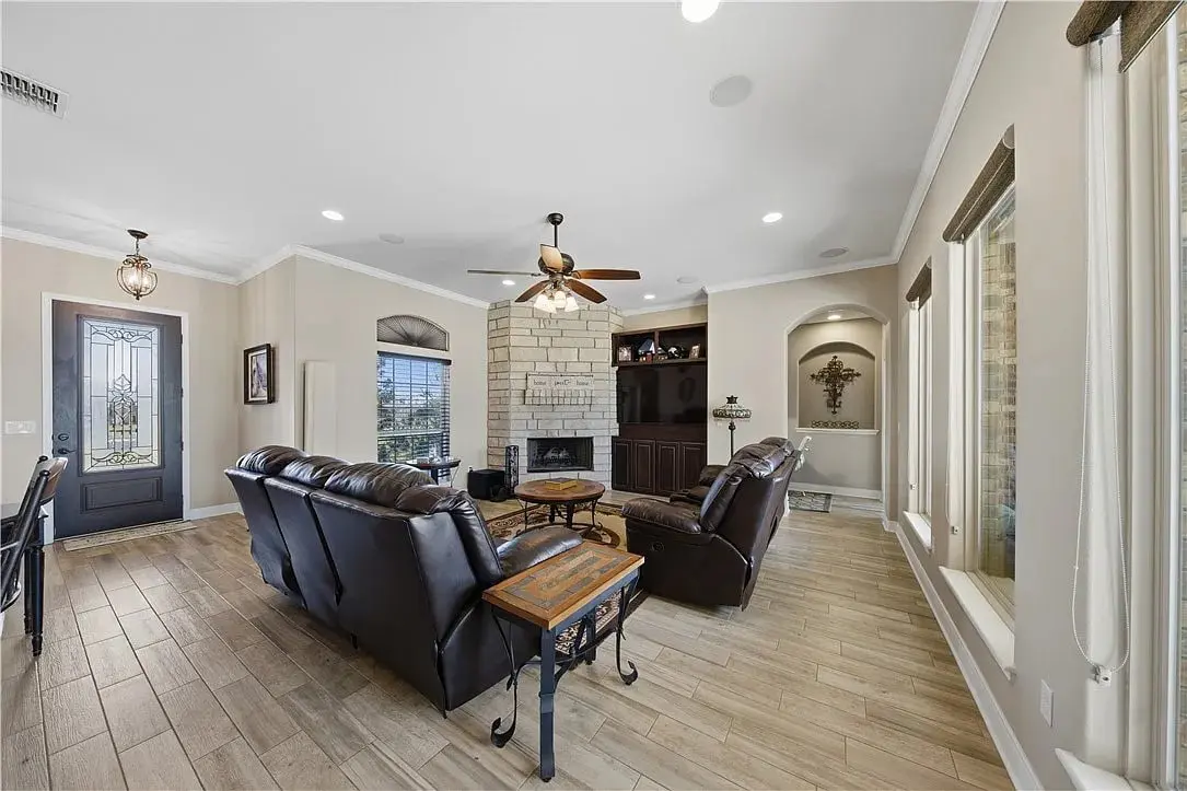 Bright living room with leather sofas, stone fireplace, built-in shelves, ceiling fan, large windows, wood-look floors.