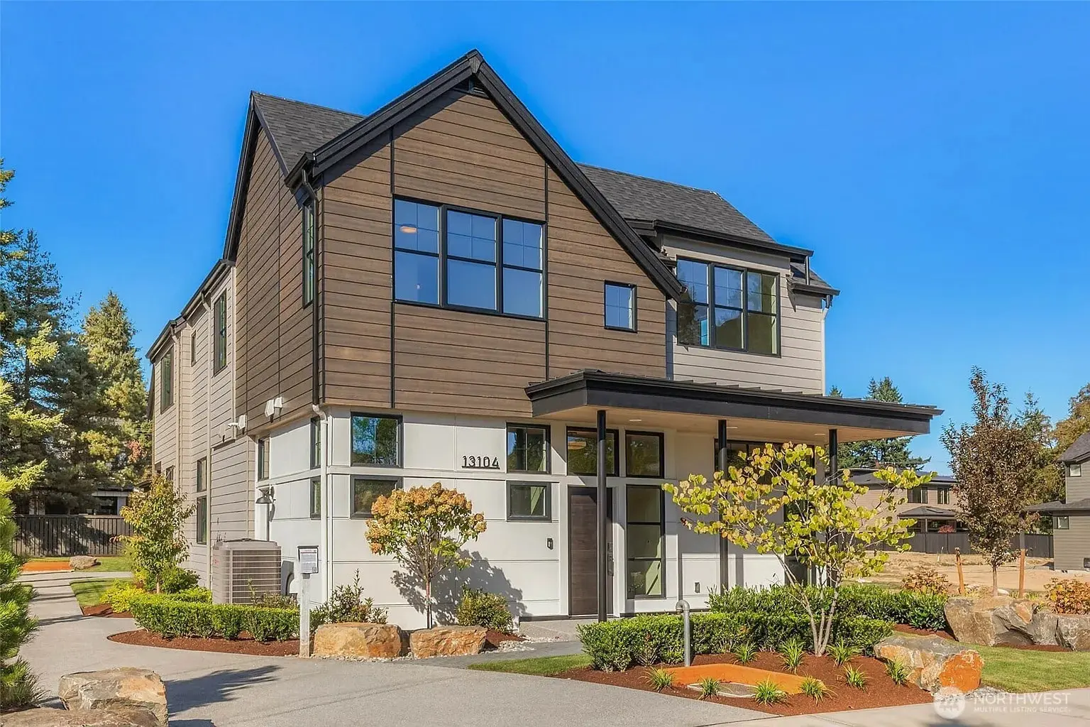 Modern two-story house with large windows, brown and white exterior, surrounded by landscaped greenery.
