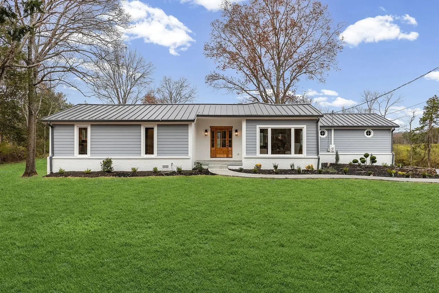 Single-story, modern gray house with a metal roof, surrounded by a green lawn and trees.
