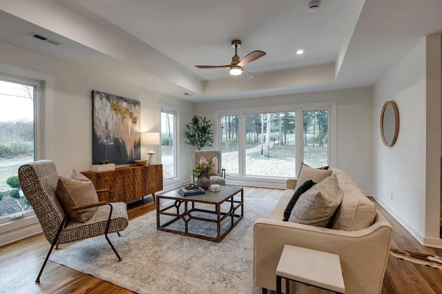 Modern living room with large windows, neutral decor, and a ceiling fan. Scenic outdoor view.