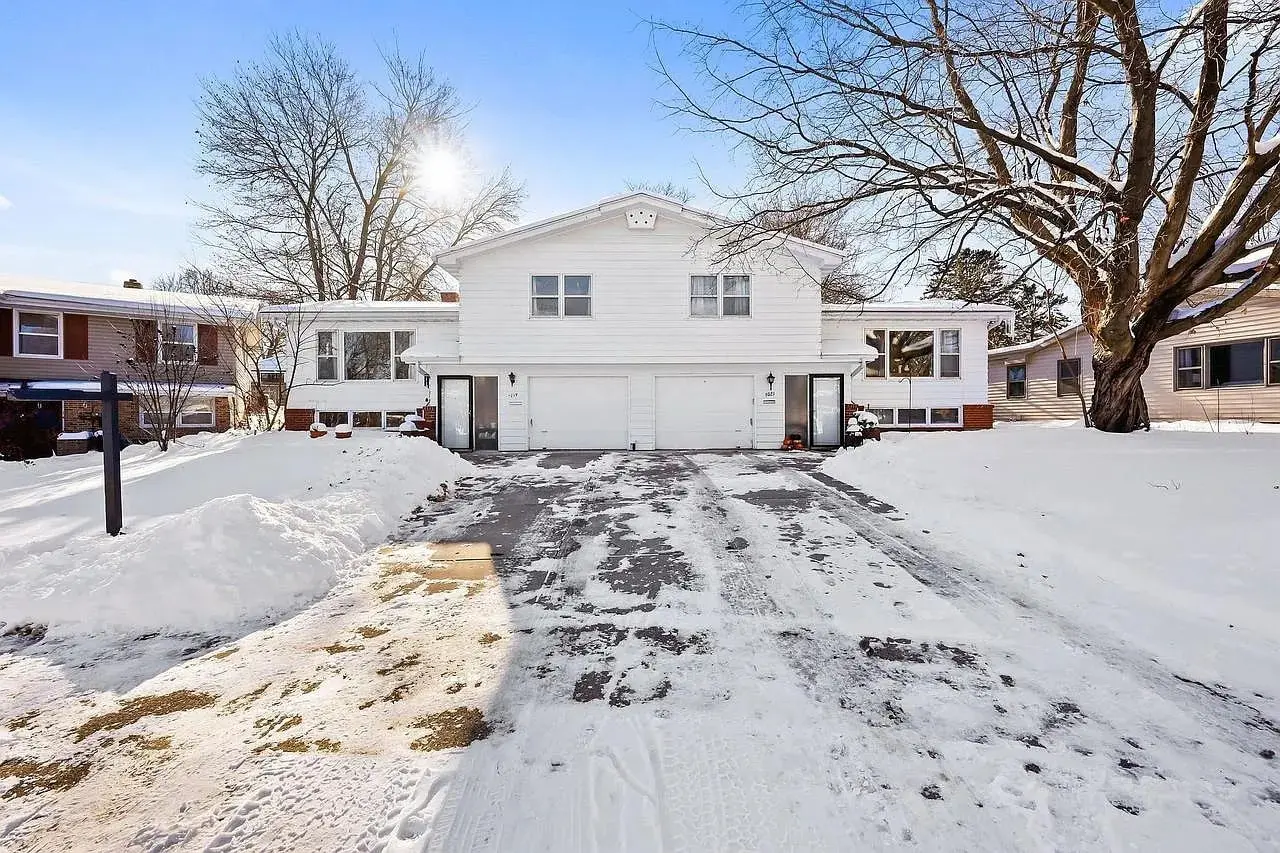 Snow-covered driveway leading to a white duplex house with a clear blue sky.