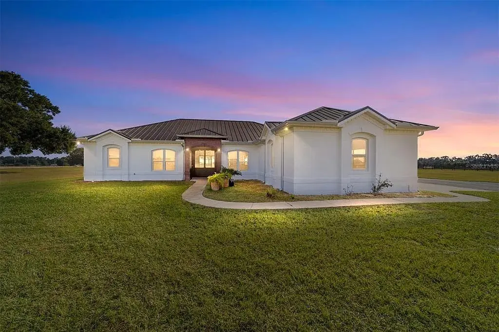 Single-story white house with a metal roof at sunset, surrounded by a grassy lawn.
