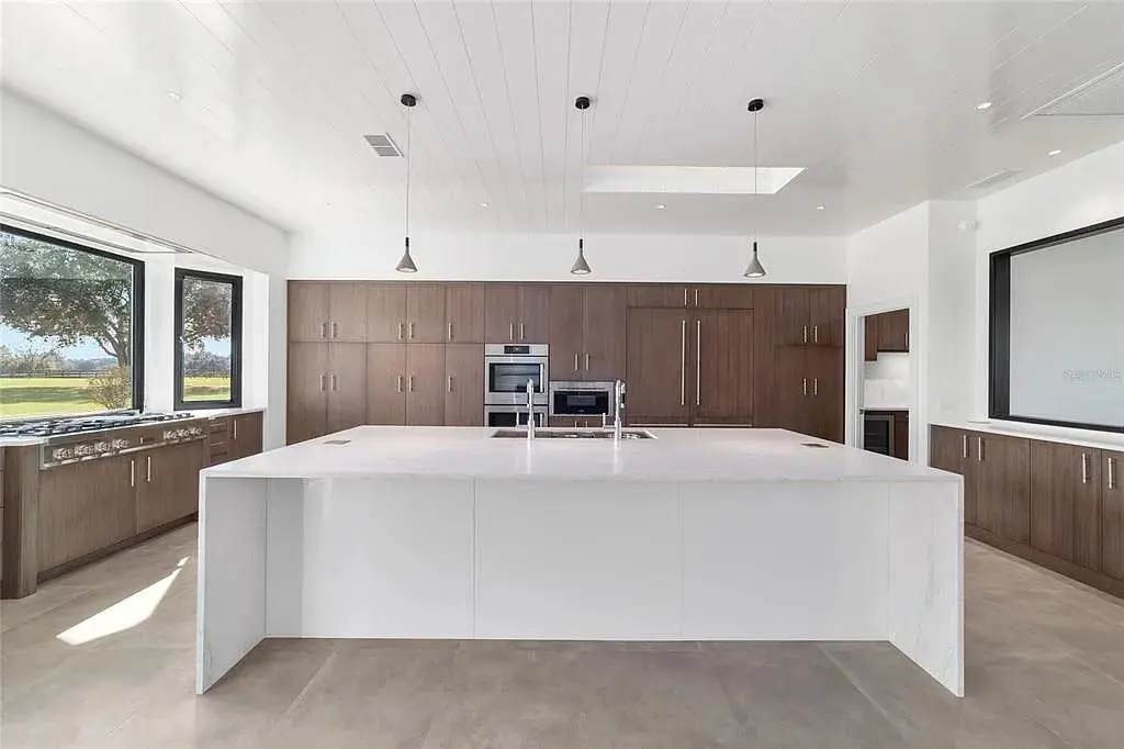 Modern kitchen with large white island, wooden cabinets, stainless steel appliances, and pendant lights.