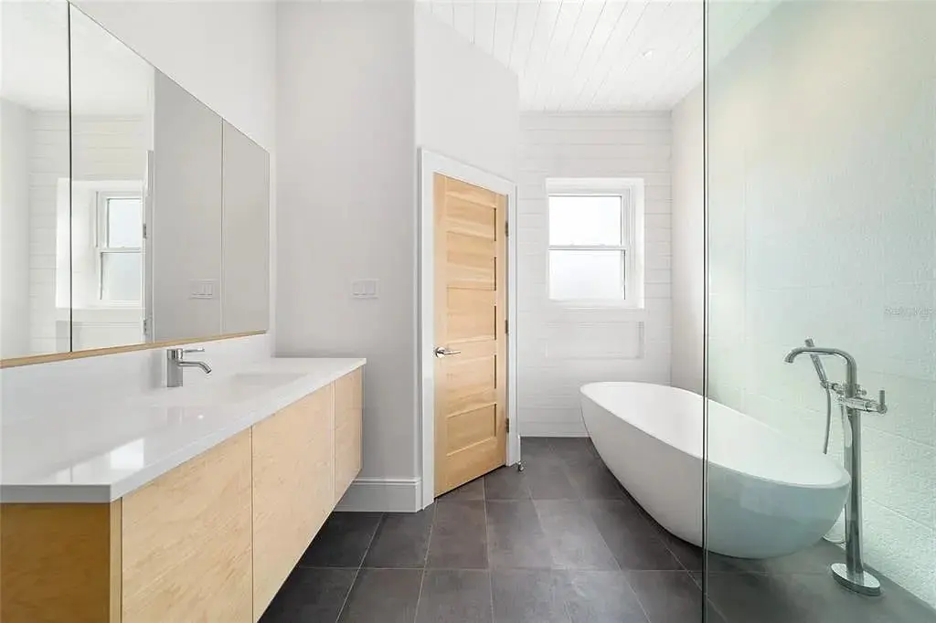 Minimalist bathroom with freestanding bathtub, wooden door, large mirror, and double sink vanity.