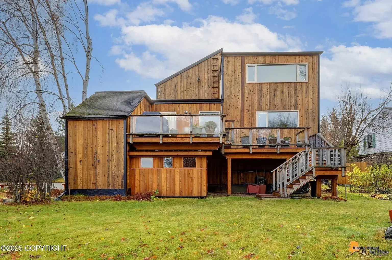 Modern wooden house with large windows, elevated deck, and surrounding green lawn under a cloudy sky.