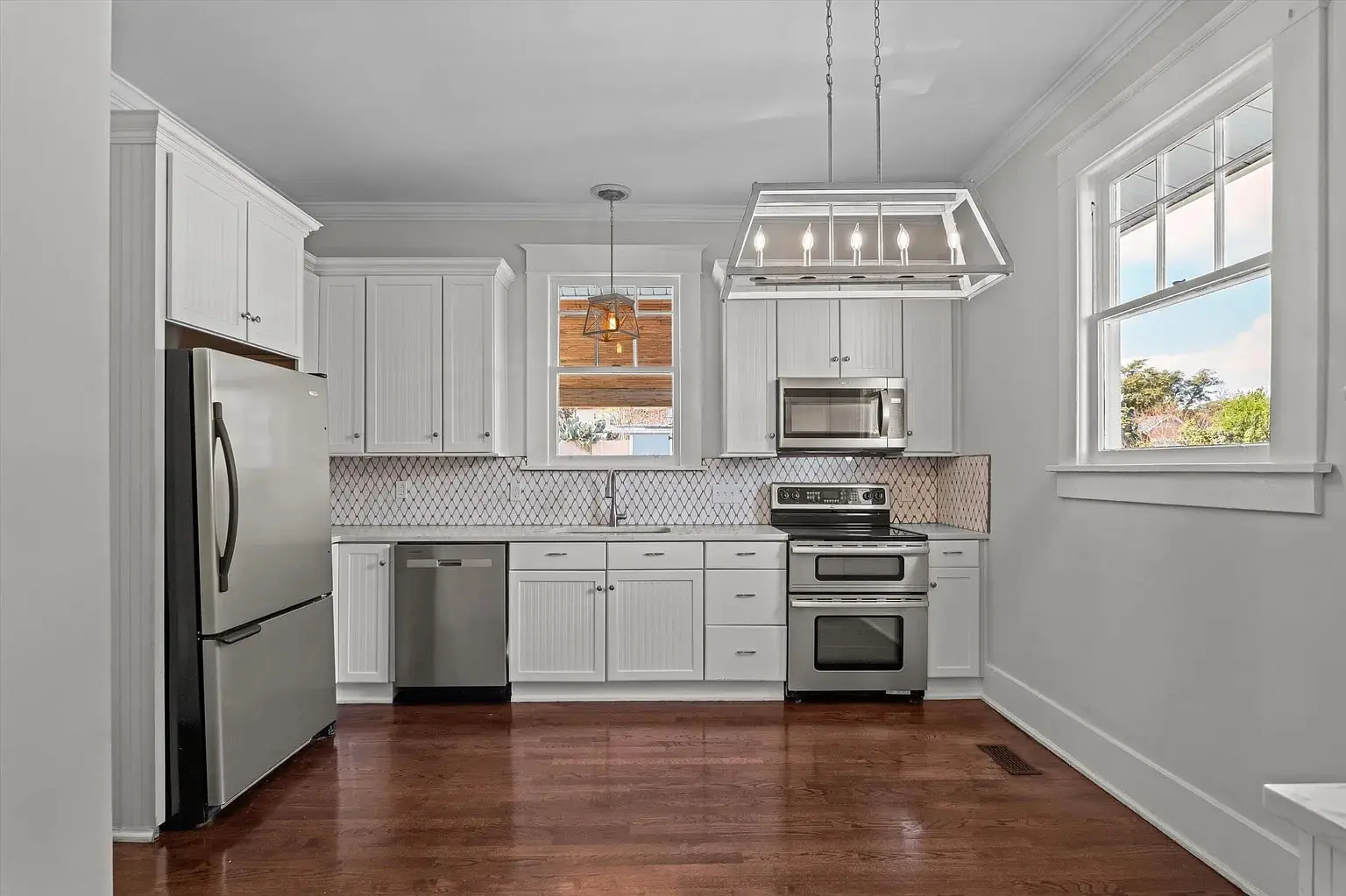 Modern kitchen with stainless steel appliances, white cabinets, and wooden floor. Large window provides natural light.