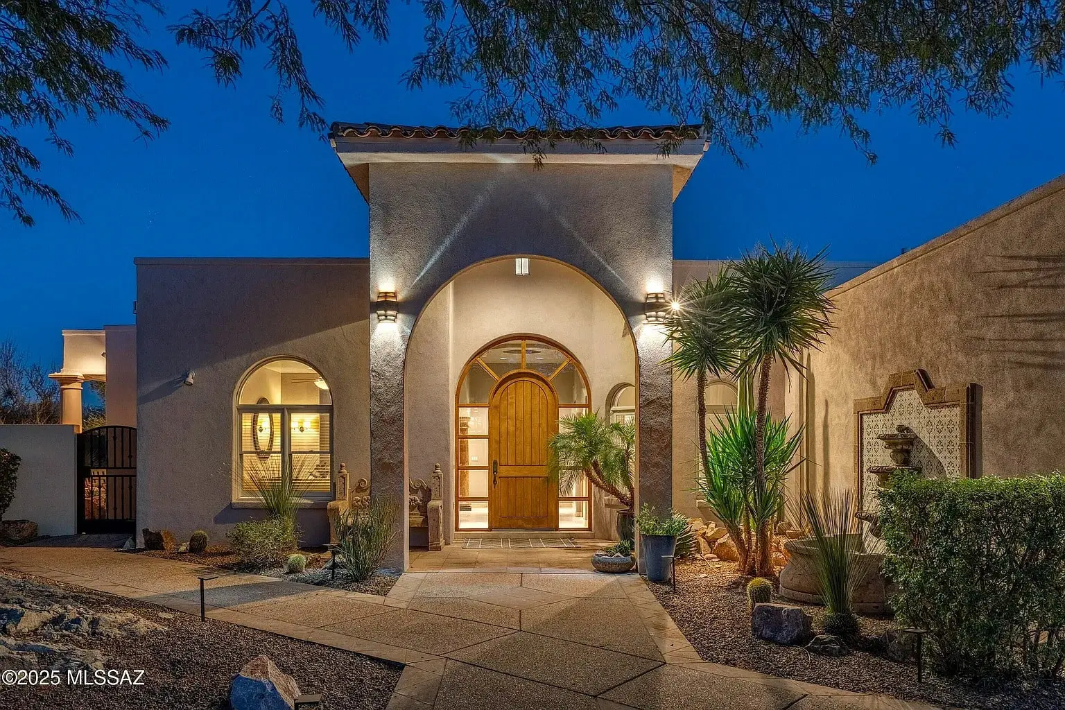 Elegant Mediterranean-style house with arched wooden door, illuminated at night, surrounded by desert landscaping.