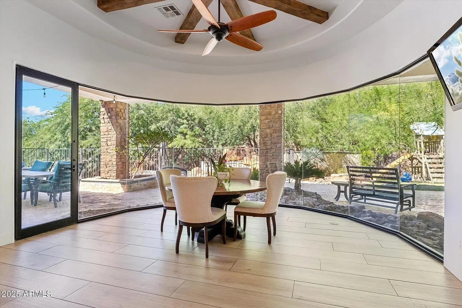 Dining area with round table and chairs, surrounded by curved glass windows overlooking a patio and garden.