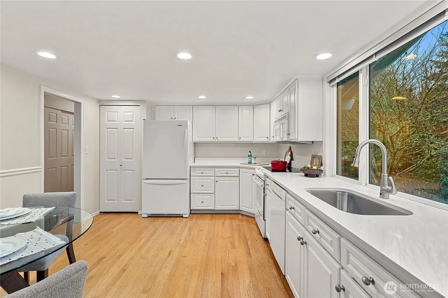 Modern kitchen with white cabinets, wooden floor, large window, and dining table with place settings.