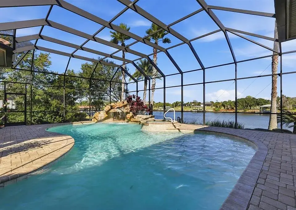 Screened-in pool area with a waterfall feature, overlooking a lake and palm trees.