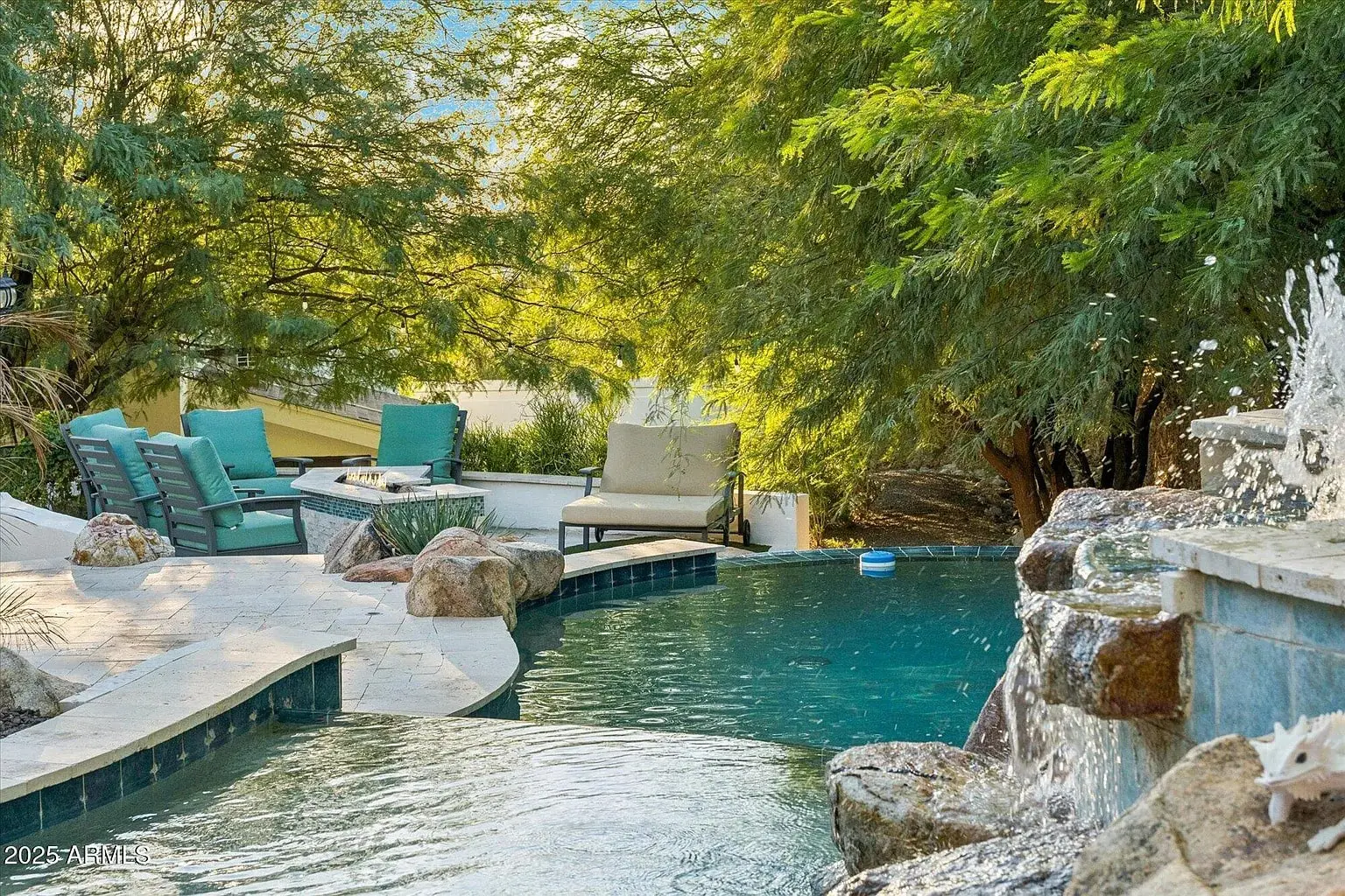 Outdoor pool area with lounge chairs, surrounded by trees and rocks, featuring a small waterfall.