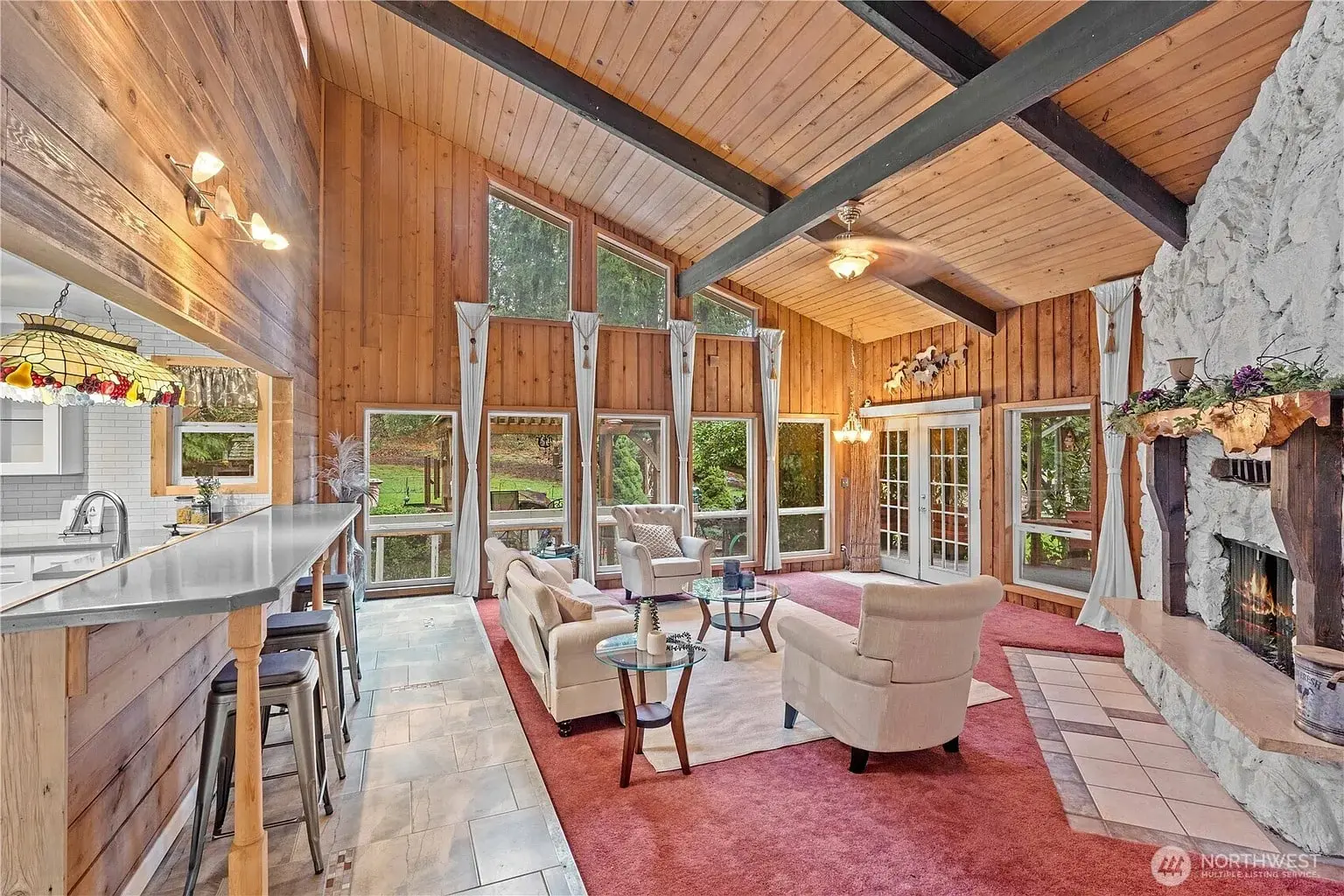 Rustic living room with vaulted wood ceiling, large windows, stone fireplace, and adjoining kitchen bar.