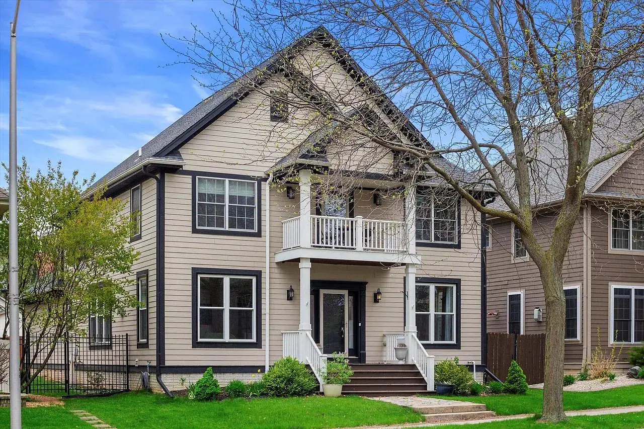 Two-story beige house with black trim, front porch, and small balcony, surrounded by a green lawn.