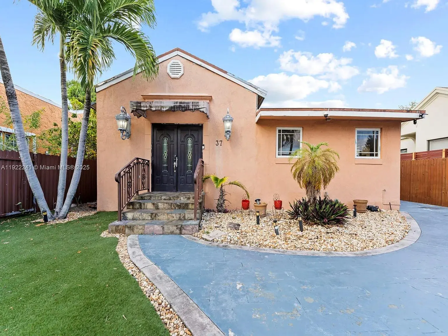 Single-story peach-colored house with a stone pathway, palm trees, and a small garden in front.