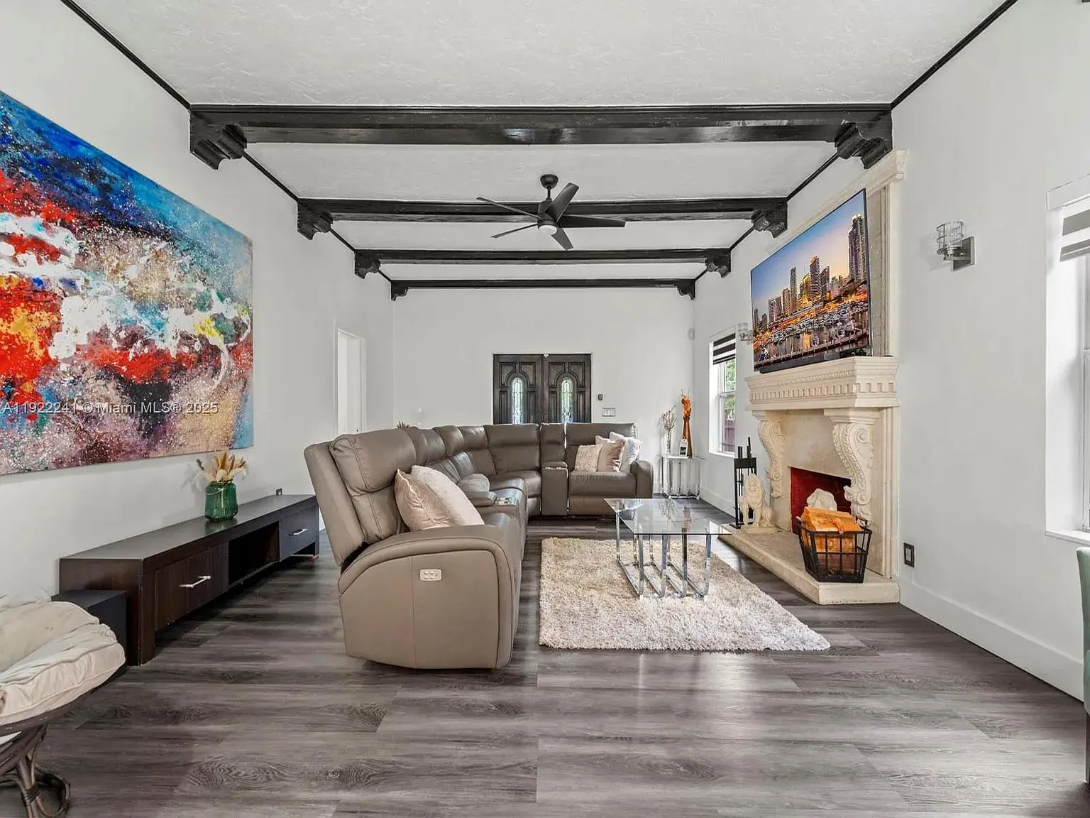 Living room with leather sectional, colorful artwork, fireplace, and dark wood ceiling beams.