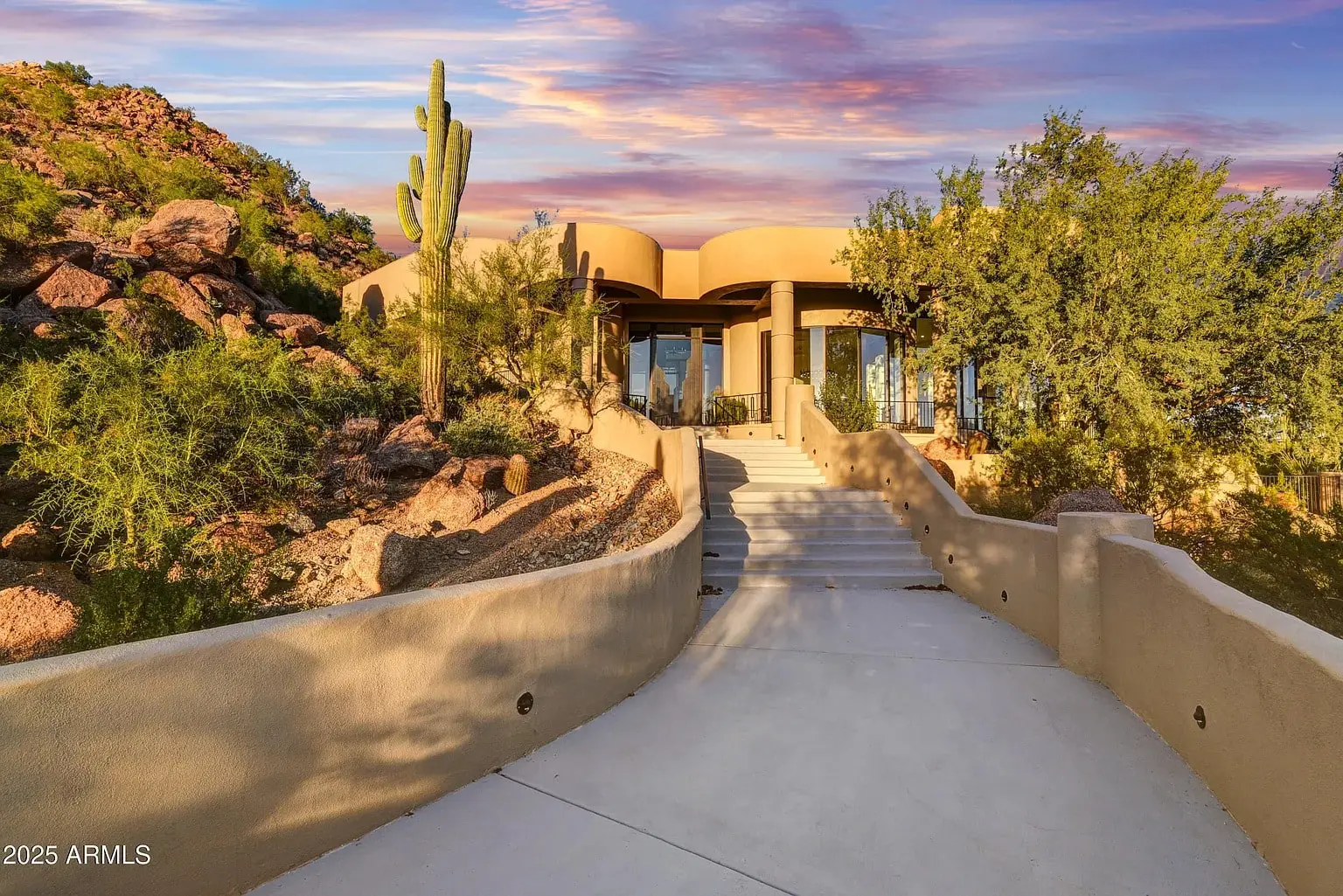 Modern desert home with large windows, surrounded by cacti and rocky landscape at sunset.