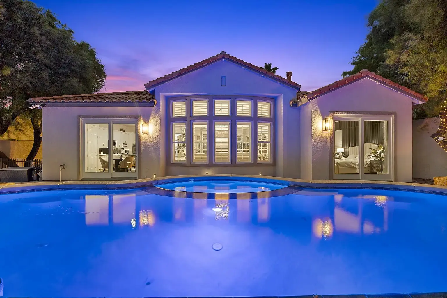 Single-story house with large windows, illuminated pool, and evening sky in the background.