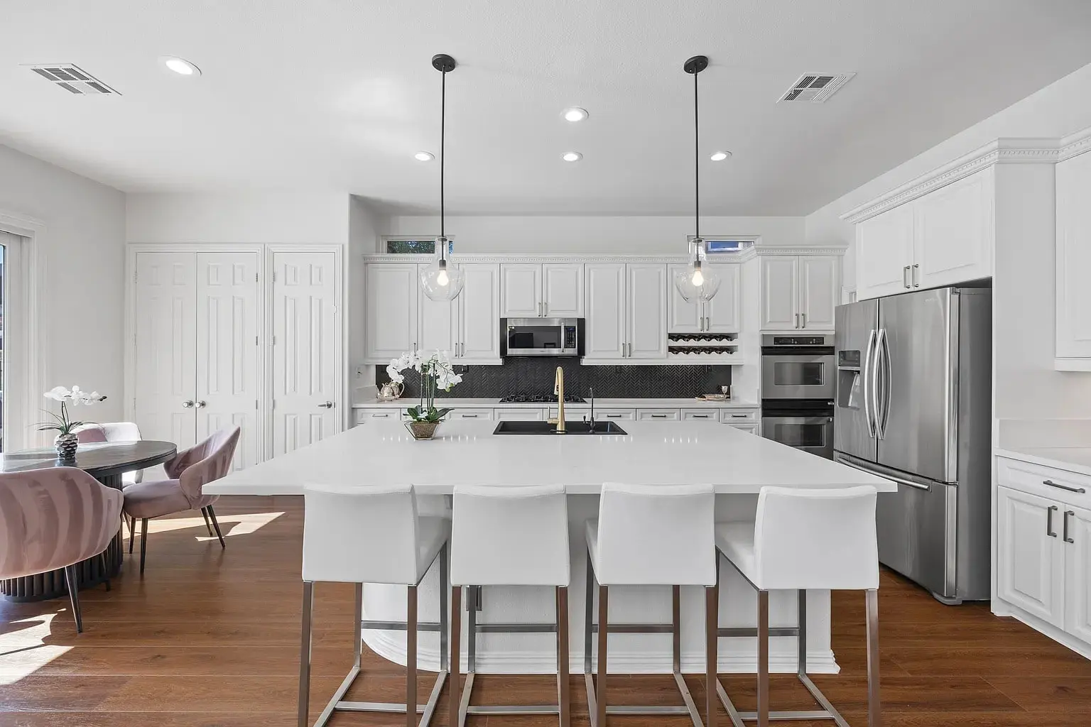 Modern kitchen with white cabinets, large island, pendant lights, and stainless steel appliances. Dining area visible.