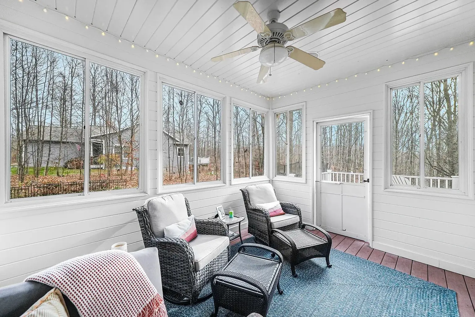 Enclosed porch with wicker chairs, large windows, ceiling fan, and view of trees outside.