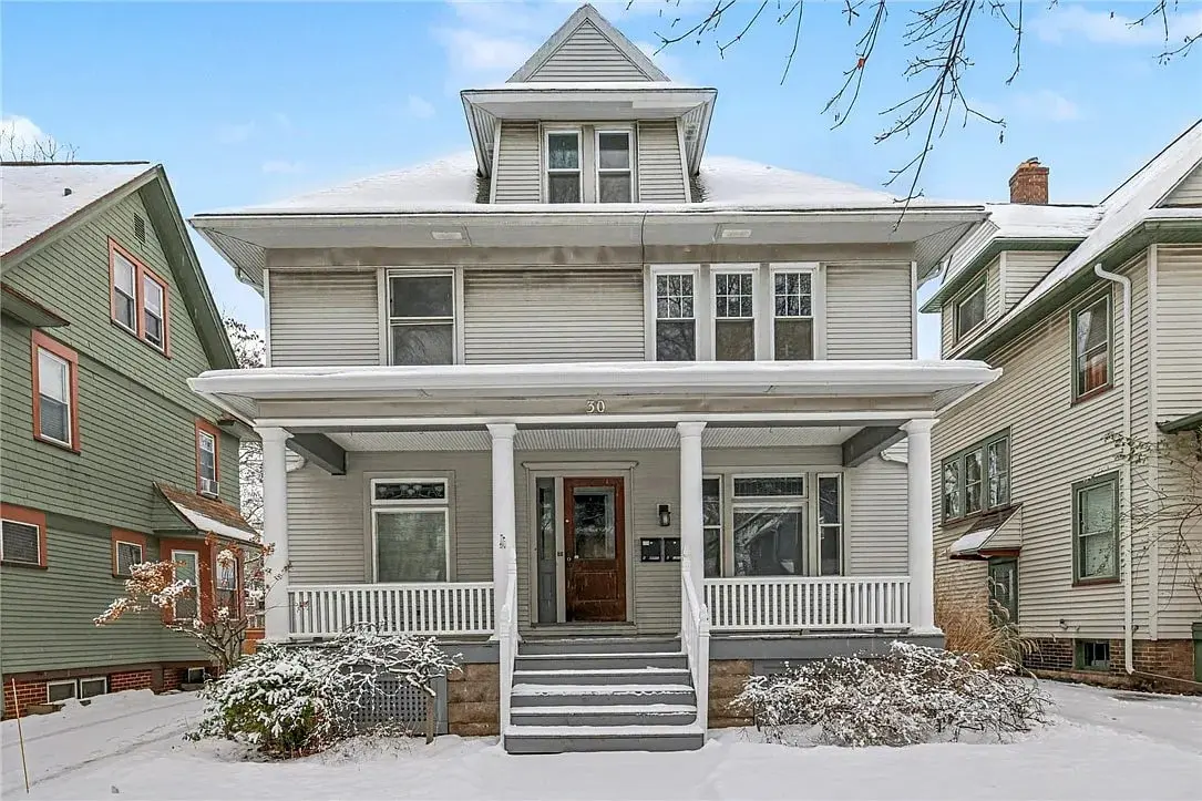 Two-story white house with a porch, surrounded by snow-covered ground and neighboring houses.