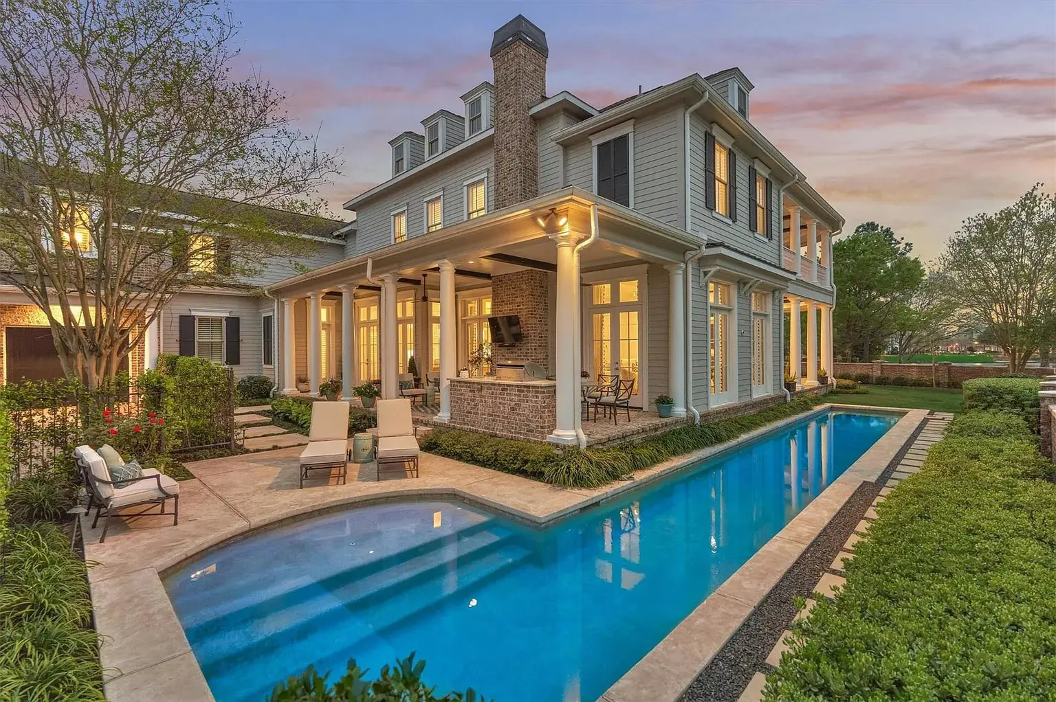 Elegant two-story house with a wraparound porch and a long, narrow swimming pool at sunset.