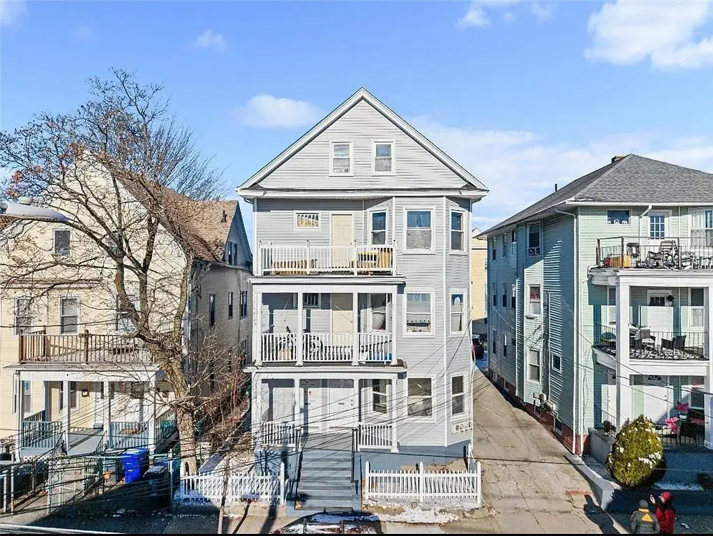 Three-story gray multi-family house with stacked porches and driveway, between similar buildings, winter scene.