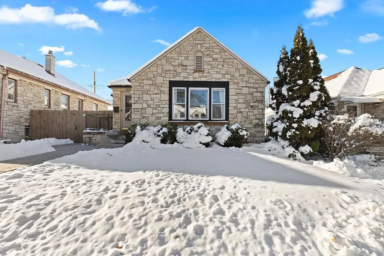 Single-story stone house with snow-covered yard and trees under a clear blue sky.