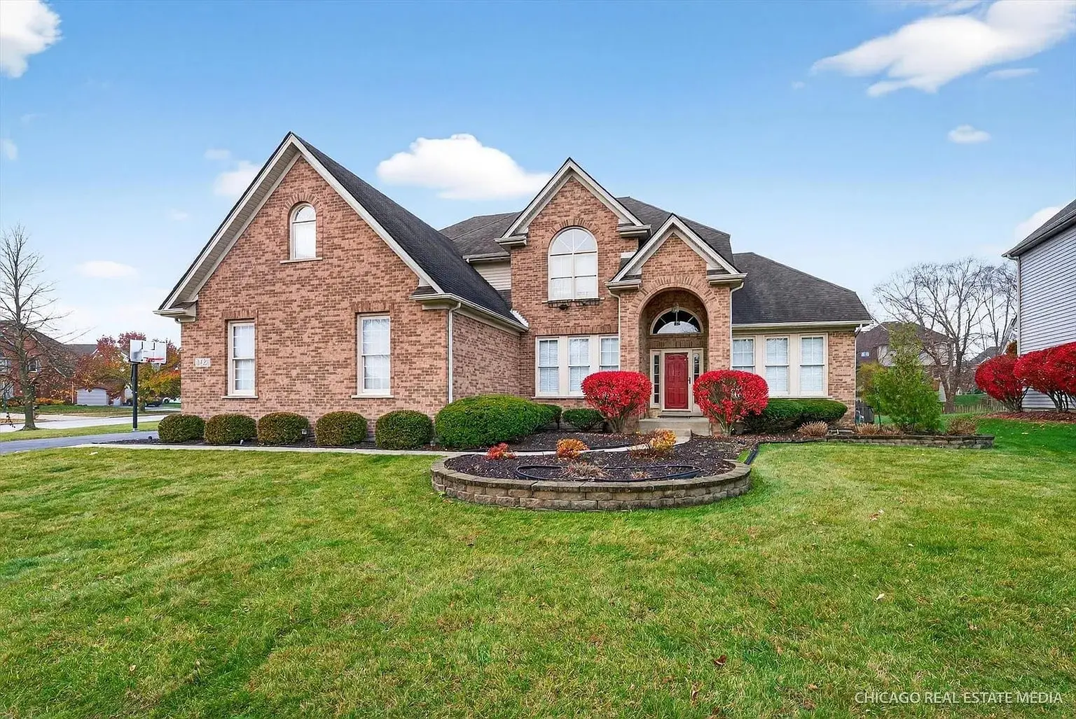 Brick house with arched windows, landscaped front yard, and red bushes under a blue sky.