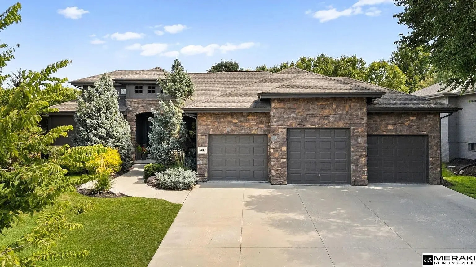 Stone-faced house with three-car garage, surrounded by landscaped greenery under a clear blue sky.