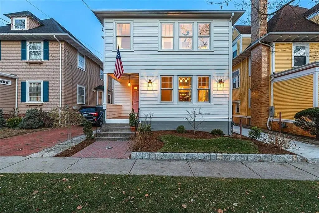 Two-story white house with an American flag, brick pathway, and front porch lights.
