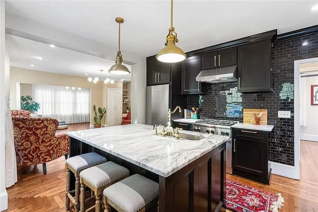 Modern kitchen with dark cabinets, marble island, pendant lights, and adjacent living area with patterned chair.