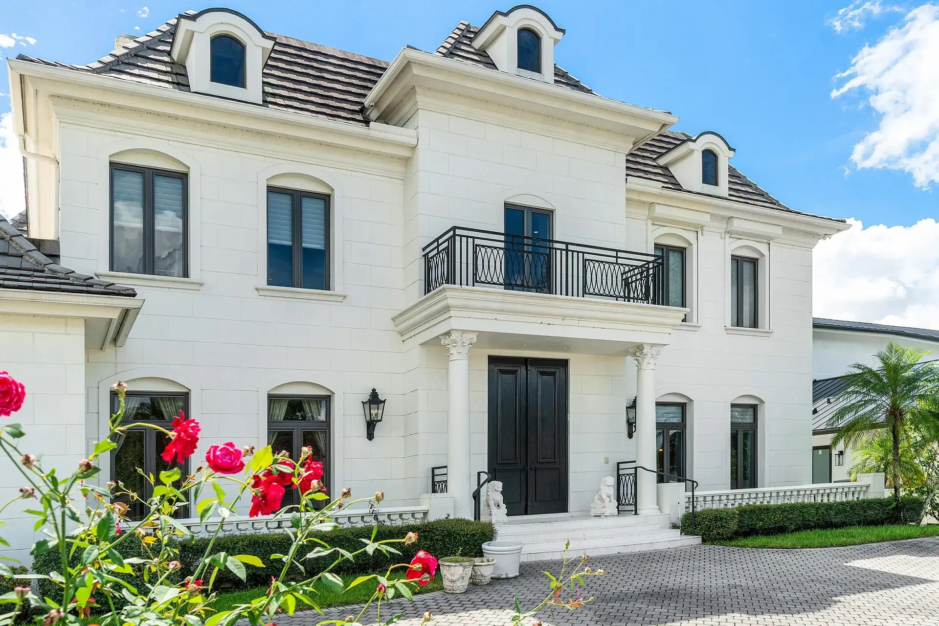 Large white mansion with a balcony, surrounded by greenery and red flowers in the foreground.