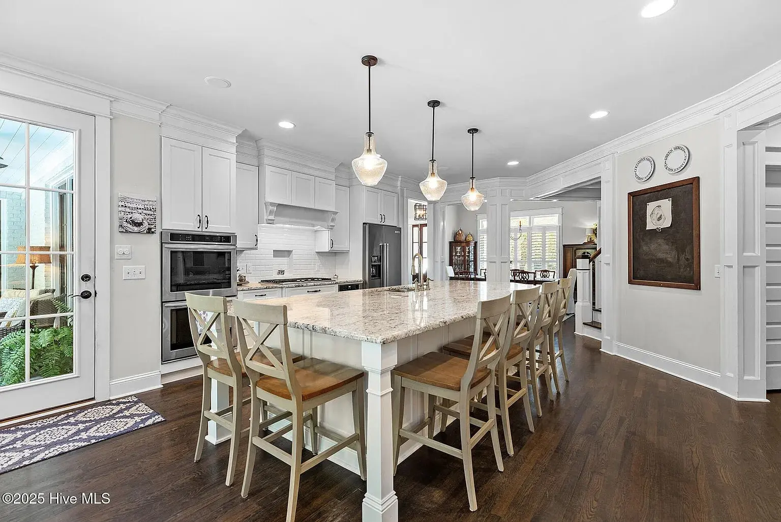 Spacious kitchen with large island, pendant lights, and wooden chairs. White cabinets and stainless steel appliances.